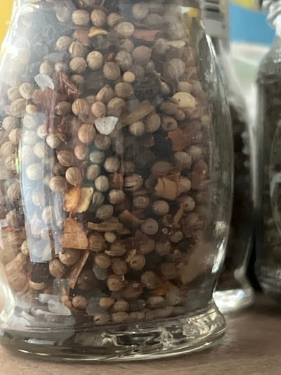 The image shows a close-up of a glass jar filled with a mixture of spices. The spices appear to be primarily whole coriander seeds, with smaller pieces of dried chili flakes and possibly other herbs or spices mixed in. Some small white crystals, which could be salt or sugar, are also visible. The jar is clear glass with a rounded base. In the background, out of focus, are other glass containers, suggesting the jar is part of a collection of spices or condiments. The lighting suggests an indoor setting. The location of Prigorodnyi, Russia is not discernible from the image content.