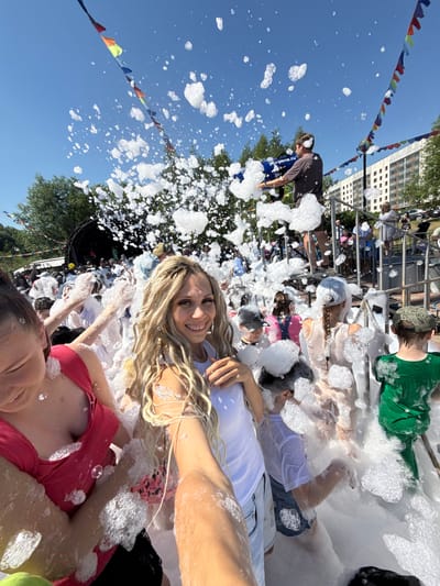 The image depicts a lively outdoor foam party in Заволжье, Russia, under a clear blue sky. A woman in the foreground, holding the camera, smiles broadly as she is covered in foam. To her left, another woman in a pink top also smiles. In the background, a person operates a foam machine, spraying a large amount of foam into the air. A crowd of people, including children, are visible enjoying the foam, some wearing hats and casual clothing. Decorative flags are strung across the sky. In the distance, buildings and trees can be seen, suggesting an urban or semi-urban setting. The ground is completely covered in a thick layer of white foam.