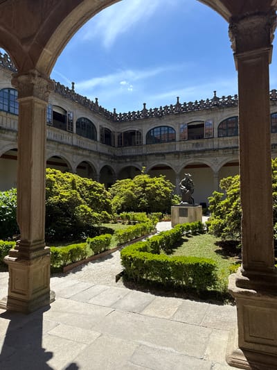 The image shows a courtyard of the Hostal dos Reis Católicos in Santiago de Compostela, Spain. The courtyard is enclosed by arcades with arched windows, and ornate balustrades line the upper story. In the center of the courtyard is a garden with manicured hedges and a statue of a figure in a robe, seated on a pedestal. The sky above is blue with scattered clouds.