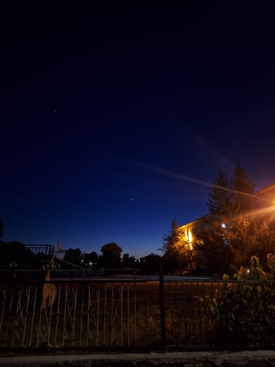 The image depicts a night scene in Dolni Chiflik, Bulgaria. A dark blue sky occupies the majority of the frame, punctuated by a few faint stars. To the right, a building is partially illuminated by a warm, yellow street light, the light also shining through the surrounding foliage. A wrought-iron fence runs along the right edge of the image, partially obscuring a road or sidewalk behind it. Bushes and trees grow on the other side of the fence, adding to the darkness of the scene. No people are visible.