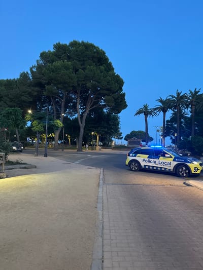A police car with flashing blue lights is parked on a street in Sant Feliu de Guíxols, Spain. The car is white with blue and yellow markings and the words "Policia Local" and "Sant Feliu de Guíxols" are visible on its side. The car is facing towards the right of the image. In the background, there are several palm trees and other trees. The sky is clear and blue, indicating it is either early morning or late evening. There is a person walking in the distance on the sidewalk. The street is paved with cobblestones, and there is a dirt path next to it. The scene appears to be in a public area, possibly near a park or promenade.