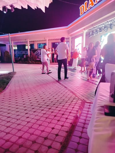 The image depicts an outdoor evening event, likely a celebration or gathering, illuminated by vibrant pink and purple lights. The setting appears to be a paved courtyard in Aksukent, Kazakhstan. In the background, a building with an illuminated sign reading "DIAMOND" is visible, suggesting the location might be a restaurant, club, or event venue.
Several people are present, engaged in various activities. In the foreground, a man in a white shirt and light-colored pants is seen dancing, his body angled as if mid-movement. Another man, also in a white shirt, stands nearby, facing him. Further back, a group of people are scattered across the paved area. One woman in a dark top and a floral skirt is walking towards the foreground, her expression suggesting amusement. Another woman in an orange top and white skirt is also dancing. Other individuals are blurred and less distinct, indicating movement or distance.
The ground is paved with a pattern of square tiles, with a contrasting darker inlay. The lighting creates a strong pink hue across the entire scene, reflecting off the pavement and the people. The overall atmosphere is lively and festive, characterized by music and social interaction.