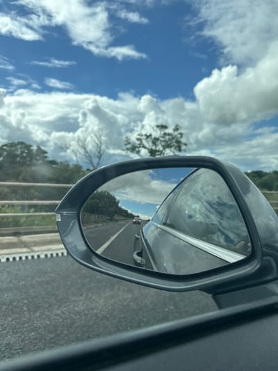 The media file shows a side-view mirror of a vehicle, reflecting a view of the road ahead and the sky. The mirror is mounted on the right side of a dark-colored car. The reflection in the mirror shows a highway with multiple lanes, bordered by green trees and foliage. A few vehicles are visible in the distance on the highway.
The sky is a prominent feature, with dramatic, fluffy white clouds scattered across a bright blue expanse. Sunlight appears to be filtering through the clouds, creating a bright and airy atmosphere. The overall impression of the sky suggests it is daytime, likely late morning or early afternoon.
The road surface is asphalt, with white lane markings visible. A concrete barrier runs along the side of the road, separating it from the adjacent landscape. A white fence or railing is also visible alongside the road, and the texture of the road surface suggests it might be slightly damp or dusty.
The reflection in the mirror also captures a sliver of the vehicle's interior, including a person's hand on the steering wheel. The perspective is from inside the car, looking out through the side window and into the side-view mirror.
The scene is consistent with driving on a highway in a rural or semi-rural area. The weather appears to be pleasant, with a mix of sun and clouds. No specific text is visible in the media file. The location context is Newry, United Kingdom.