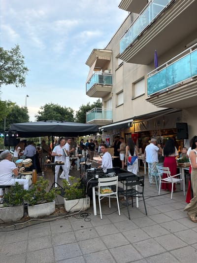 The image depicts a live musical performance at a restaurant/bar, likely in Cambrils, Spain, based on the presence of Cuban flags and the style of architecture. Several musicians are performing on the sidewalk outside the establishment, positioned under a black canopy. Most musicians are wearing white outfits and playing various instruments like drums, percussion, and potentially keyboards. A small crowd of patrons is seated at tables, watching the performance. Some are inside the covered area of the restaurant and some sit outside in chairs on the sidewalk. The building next to the restaurant has modern balconies and a blue awning. The sky is partly cloudy and trees line the street. The ground is paved with stone tiles. Plant pots with greenery are visible. Electrical wires are present on the ground.