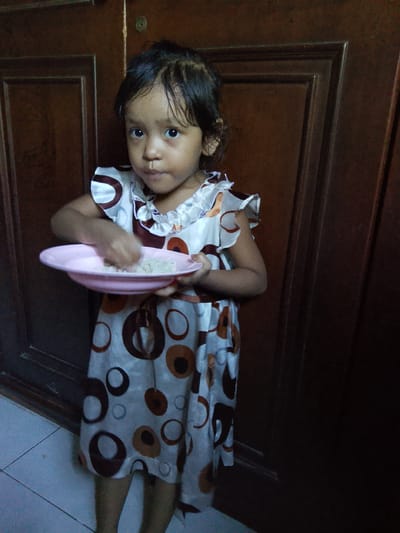 In Merbopuntong, Indonesia, a young girl stands in front of a dark wooden door. She is wearing a dress with a pattern of brown and white circles. The girl is holding a pink plate in her left hand, and her right hand is in the plate, seemingly mixing or eating its contents, which appear to be rice. The floor in front of her is tiled.