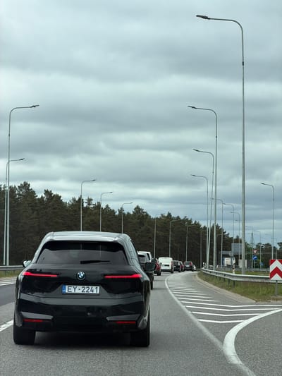 The image shows a black BMW car with license plate EY-2241 on a highway. The car is likely in a traffic jam, as other vehicles are visible ahead in the same lane. There is a forest area alongside the road, and a series of tall streetlights line the shoulder of the road. A sign indicating a curve in the road is visible. The sky is overcast with dark clouds. The scene is in Berģi, Latvia, based on the license plate country code "LV" for Latvia.