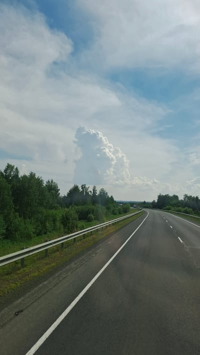 The image shows a highway in a rural area. The road is asphalt and has white lines marking the lanes. There is a metal guardrail along the left side of the road, and beyond that, a dense green forest. The sky is partly cloudy with a large, dramatic cumulonimbus cloud formation on the left. The time of day appears to be daytime. There are no people visible in the image. The image is taken from a vehicle moving along the highway.