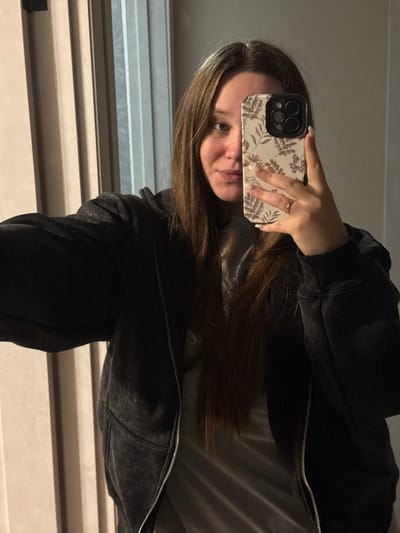 A single female is depicted in a mirror selfie, extending her left arm toward the camera. She possesses long, dark brown hair, which falls over her shoulders, and a light complexion. Her left eye is visible, displaying a blue-green hue, while her lips are slightly parted. She is attired in a dark gray, washed-out zippered hoodie, worn open over a lighter gray t-shirt. Her right hand, adorned with a thin gold-colored ring on the ring finger, holds a smartphone. The phone features a light-colored case patterned with dark botanical designs, including leaves and small butterflies, and partially obscures the right side of her face. The background consists of light-colored vertical panels or blinds. This scene occurs indoors in the city of Tlustenkhabl, Russia.