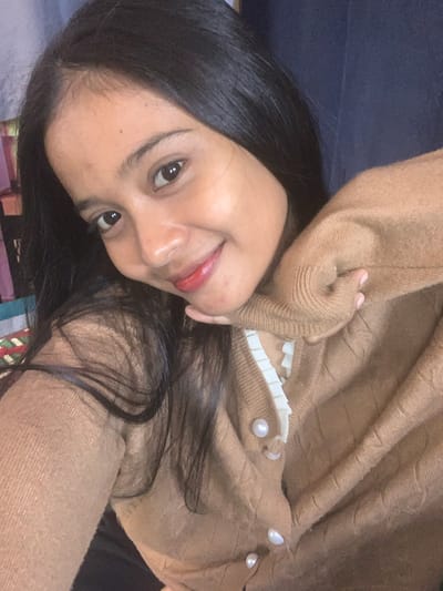 The image is a close-up selfie of a young woman with long, dark hair and brown eyes. She is wearing a light brown, cable-knit cardigan with pearl buttons and a white ruffled collar peeking out from underneath. Her right hand is resting on her chin, and she is smiling at the camera. The background is out of focus and appears to be an indoor setting with some colorful objects visible on the left side. The woman's skin has a warm tone, and she is wearing red lipstick. The lighting suggests an indoor environment, possibly with natural light. There is no discernible information in the image that can be used to determine its location in Sungai Meriam, Indonesia.