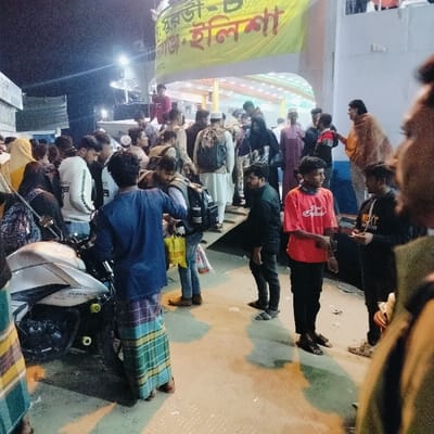 The image displays a crowded night scene at a ferry terminal in Bhola, Bangladesh, specifically a location serving the Hulisha route. Approximately 25-30 individuals, predominantly adult males with a few females, are gathered on a concrete or paved surface illuminated by artificial lights. Many individuals are carrying backpacks and bags, indicative of travel. In the mid-left, a light-colored motorcycle is parked. The background features a white structure with blue accents, likely a ferry vessel or terminal building, above which a yellow banner with Bengali text "Purabi-6 / Dauran • Hulisha" is prominently displayed. People are standing, congregating, and appearing to wait or move towards the structure. Essential figures include a male in a blue shirt and multi-colored plaid lungi next to the motorcycle, a young male in a red t-shirt, and another young male in a black hoodie holding a smartphone. Scattered debris is visible on the ground.