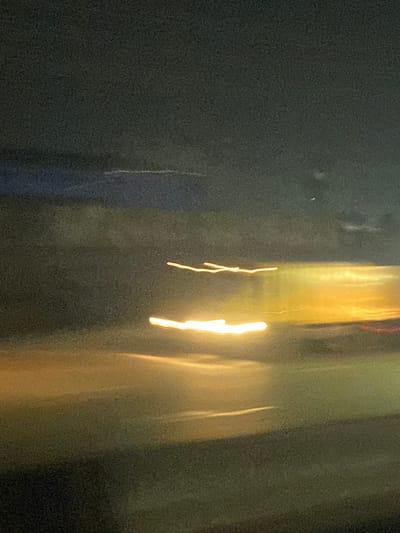 A blurry, abstract image. On the left, a dark, textured area. Moving right, vertical streaks of light yellow and white appear, resembling light trails. The background shifts from dark to murky with undefined shapes. It's difficult to discern concrete objects due to the blur, suggesting movement or an out-of-focus shot at night.
