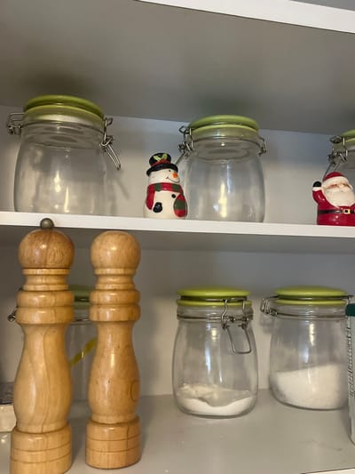 The image displays the interior of a white cabinet or shelving unit, likely in a domestic setting within Bogota, Capital District, Colombia. The scene is static and well-lit.

On the upper shelf, three clear glass jars with light green, hinged clamp-style lids are visible. The leftmost and middle jars appear empty, while the rightmost jar is partially obscured and also empty. Positioned between the leftmost and middle jars is a small, white ceramic snowman figurine, adorned with a black top hat, green holly, a red and green striped scarf, and featuring black dots for eyes and a small orange-brown nose. To the right of the middle jar, a red and white ceramic Santa Claus figurine is partially visible.

The lower shelf holds two tall, light-brown wooden grinders, consistent with salt and pepper mills, positioned on the left side of the shelf. To their right, two additional clear glass jars with light green, hinged clamp-style lids are present. The left jar on this lower shelf contains a small quantity of white powdered substance. The right jar on this lower shelf contains a significant amount of white granulated substance. A vertical white object is partially visible on the far right edge of the lower shelf.