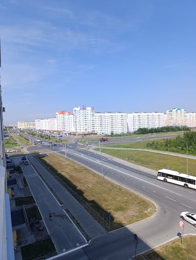 The image shows a wide, modern avenue in Nizhnevartovsk, Russia, bordered by numerous residential buildings on both sides. The sky is clear and blue with sparse clouds. On the left side of the avenue, there are multi-story apartment buildings, some with colorful accents. A sidewalk runs alongside these buildings, with a few people walking and a group of parked bicycles. Further down the avenue, there is a traffic light and a crosswalk.
On the right side of the avenue, more residential buildings are visible, stretching into the distance. These buildings are also multi-story and appear to be modern in design. The avenue itself has multiple lanes of traffic. A white bus is driving on the right side of the road. A white car is parked on the side of the road near the right edge of the image. Green grass and trees line the median and the sides of the avenue, indicating a well-maintained urban environment. The overall impression is a typical scene of a residential district in a Russian city.