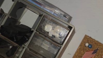 The image shows a section of a grey plastic storage box with multiple compartments. One compartment contains several white, round objects that appear to be lenses or small discs, some possibly packaged in clear plastic. Another compartment seems to contain a dark, possibly electrical, object. In the background, there is a corner of a corkboard with a miniature American flag and two small, round metal or plastic beads. The photo was taken indoors, likely on a white surface. There is no contextual information in the image to indicate its location in Lompoc, United States.