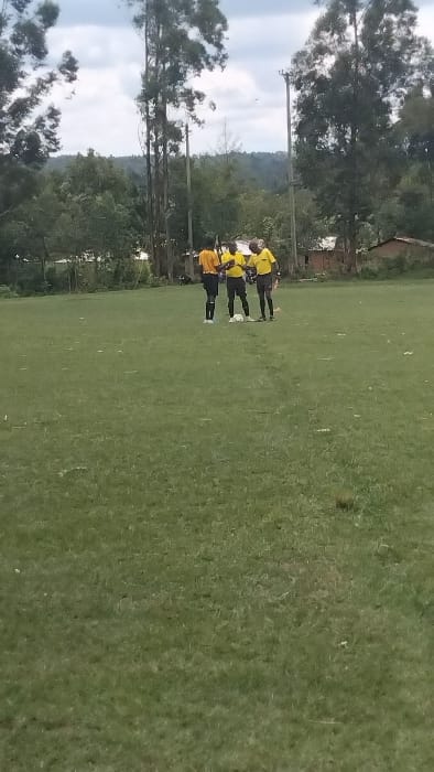 The image shows three men in yellow shirts and black shorts standing on a grassy field. The field is surrounded by trees and shrubs. The men are interacting with each other, possibly discussing the game. The location is a soccer field in Bungoma, Kenya, at coordinates 0.554 latitude, 34.774 longitude. The sky is overcast with a light grey hue. There is a slight breeze blowing through the grass. The image was likely captured during a soccer match.
