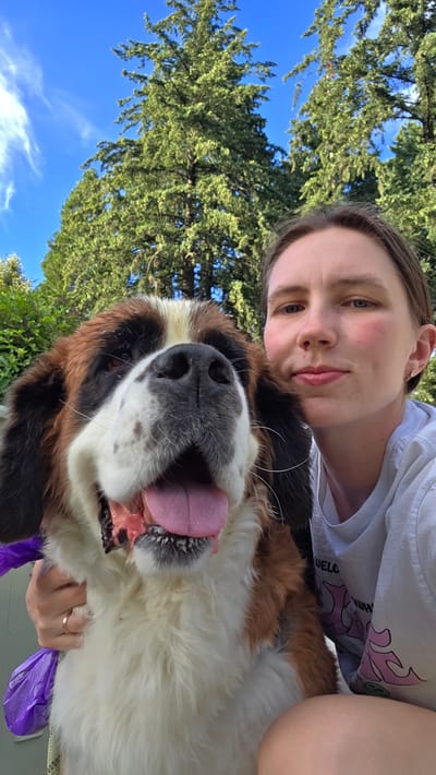 A young Caucasian woman is taking a selfie with a large St. Bernard dog. She is in the foreground, to the right, with her head tilted and a faint smile. The dog is in the left foreground, with its mouth open and tongue out, looking directly at the camera. Both are outdoors, with tall evergreen trees and a bright blue sky with wispy clouds in the background. The woman is wearing a grey t-shirt. The dog has brown, white, and black fur. The setting appears to be a park or backyard in Portland, Oregon, United States of America.