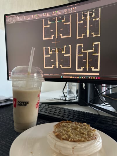 The image shows a workspace, likely in an office or home office environment. In the foreground are a monitor displaying what appears to be an architectural floor plan design in AutoCAD, a beverage from "Coffee House" in a plastic cup with a straw, and a piece of cake on a white plate. A black computer keyboard is also present. The floor plan shows room layouts, potentially of apartments or housing units, labeled with terms like "BALCONE" and "CAMERA" (likely meaning bedroom). The beverage has a light brown color. The cake has white frosting and a textured top, possibly consisting of nuts. The location in Zovuni, Armenia, is not discernible from the visual information in the image.