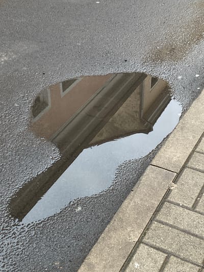 The image shows a puddle on an asphalt surface, reflecting a building. The puddle is situated next to a sidewalk made of interlocking pavers, with a concrete curb separating the asphalt from the pavers. The reflection in the puddle shows the upper portion of a building with windows and a reddish-brown facade, and a sloped roofline. The asphalt surface is wet and textured, with small ripples visible on its surface near the edge of the puddle. The lighting suggests it is daytime, with shadows visible on the paved area. The scene is a common urban environment, likely a street or sidewalk in Bílina, Czechia. No people or animals are visible in the image.