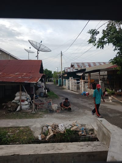 The image depicts a street scene in Geudonggeudong, Indonesia, likely during the daytime given the lighting. The street is paved with asphalt and bordered by residential buildings and vegetation.

In the foreground, a concrete structure and a drainage ditch are visible. Piles of sacks, some appearing to contain cement or construction materials, are stacked near a dilapidated shed with a corrugated metal roof. A wheelbarrow, a shovel, and several buckets are also present, suggesting ongoing construction or maintenance work.

Two individuals are engaged in activities on the street. One person, wearing a blue shirt and red cap, walks away from the camera towards the right. Another person, crouched down, is working with materials in buckets near the left side of the street.

Further down the street, more houses with corrugated metal roofs can be seen, along with satellite dishes mounted on some of the structures. A white car is parked in front of a house with an ornate metal gate. Power lines crisscross the sky above the scene. A mature tree with dense green foliage is visible on the right side of the image. The sky is overcast.