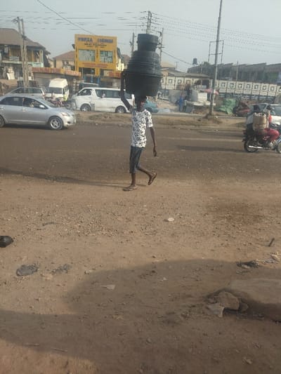 A man in a white shirt with black patterns is walking down a dirt road. He is carrying a large, black, plastic container on his head. The road is in the middle of a town, and several buildings are in the background. There are several cars parked in the background, as well as some people walking. In the far right corner of the picture, a man on a motorbike is driving away from the camera. The scene is in Way, Nigeria, at coordinates (latitude: 9.029, longitude: 7.584).