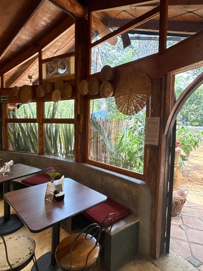 The image shows an interior view of a building with a wooden structure, possibly a restaurant in Tetela Del Monte, Mexico, as suggested by the surroundings. There is a table setting visible with a wooden table, chairs, and a table caddy with flowers. The walls and ceiling have exposed wooden beams and framed windows offering views of lush greenery, suggesting a garden or natural environment outside. Several decorative woven baskets are attached to the walls. Natural light is streaming in from the windows. There is a sign on the wall above the window, possibly with some regulations or information. Outside, there appears to be a bag and some containers of baskets. The building materials include wood, stone, and possibly concrete.