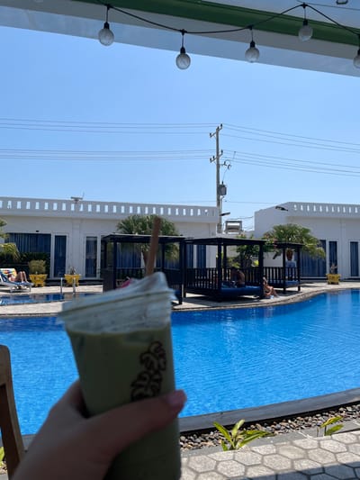 A person holds a green beverage with a straw in the foreground. The background features a swimming pool with blue water and sunbathers on lounge chairs. White buildings with palm trees and black railings line the pool. Overhead power lines and a light pole are visible in the sky. The image is taken on a sunny day.