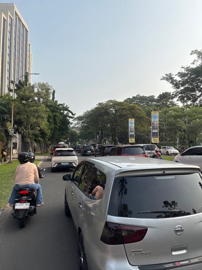 The image depicts a traffic jam on a street in South Tangerang, Indonesia. The scene is dominated by vehicles, including cars and motorcycles, all stationary or moving slowly. In the foreground, a silver station wagon is prominent, its rear end facing the viewer. To the left of the station wagon, a motorcycle with two occupants is seen from behind, with the rider wearing a light-colored shirt and blue jeans, and the passenger in a light pink shirt and blue jeans. The motorcycle's license plate is visible, reading "B 4592 UEG".
Further down the street, a white sedan with license plate "B 1803 BDS" is visible. On the right side of the road, there are several parked cars and a white Volkswagen Beetle. The street is lined with trees and buildings, including a tall, modern building with many windows on the left. Two large banners promoting a housing development are displayed on poles amidst the trees. The sky is clear and bright, suggesting it is daytime. The overall impression is one of urban congestion and daily life in an Indonesian city.