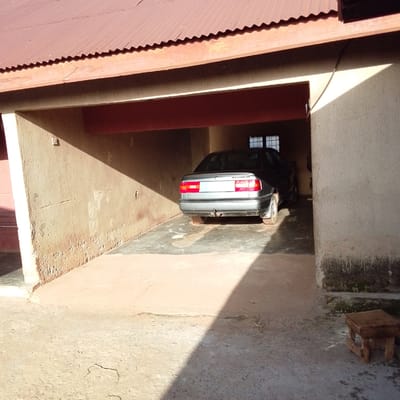This media file captures a view of a residential or commercial building's exterior, possibly a carport or garage area, under a corrugated iron roof. The scene is outdoors, with natural sunlight casting strong shadows, suggesting it is daytime. The walls appear to be made of concrete or a similar material, with some signs of wear and tear, including cracks and peeling paint.
The primary object in the scene is a silver sedan parked within the covered area. The car is facing away from the viewer, showing its rear, including taillights, bumper, and a trailer hitch. Through an opening in the structure, a glimpse of a window with multiple panes can be seen inside the building.
In the foreground, to the right of the image, is a small, rustic wooden stool with four legs. The ground is a mixture of concrete and dirt, with some areas appearing damp or stained. A coiled rope or similar object is visible on the ground in the foreground.
The overall impression is that of a functional outdoor space. The time of day is clearly indicated by the sharp shadows. The location cues, such as the architectural style and the general appearance of the surroundings, are consistent with a tropical or sub-tropical climate. There is no visible text in the image. The scene is devoid of people, focusing solely on the setting and the parked vehicle.