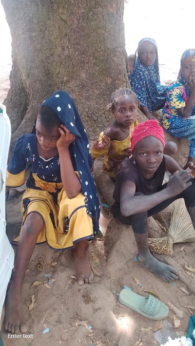 This image depicts five young females, appearing to be children or adolescents, seated on dry, dusty earth at the base of a large tree with textured bark and visible roots in Rinze, Nigeria. The scene is illuminated by natural light, indicating daytime.
On the far left, a female child wears a dark blue dress with yellow long sleeves, a yellow skirt, and yellow floral appliqués. A dark blue head covering with white polka dots is draped over her head and shoulder. She looks downwards and to her left, with her left hand raised to her temple. Her bare feet rest on the soil, which appears dusty.
To her right, a smaller female child sits behind her, wearing a yellow patterned top, with hair styled in small braids. She looks directly towards the viewer. Behind her, another female child is partially visible, wearing a blue garment with a white floral pattern and a blue head covering, also looking towards the viewer.
In the foreground on the right, a female child wears a red headscarf, a dark top, and dark leggings. She looks directly at the viewer. Her bare feet and lower legs are noticeably covered in a dark substance, consistent with dry earth or dust. A single light green flip-flop sandal and a bundle of dried, light-colored straw (resembling a broom) lie on the ground near her feet.
A fifth female child is partially visible on the far right, wearing a blue garment with yellow and orange patterns. Her face is mostly obscured. Small pieces of light blue debris are scattered across the dusty ground.