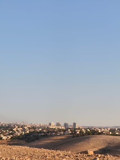 The image shows a panoramic view of the city of Keidar, located in the Palestinian Territories. In the foreground, a rocky, barren hillside is visible, with sparse vegetation and a clear blue sky overhead. The middle ground features a sprawling residential area with numerous houses, many of which have red-tiled roofs, interspersed with trees and greenery. Further in the background, a denser urban landscape emerges, characterized by taller buildings and a hazy skyline. The sun appears to be low in the sky, casting long shadows and bathing the scene in a warm, golden light. There are no people visible in the image.