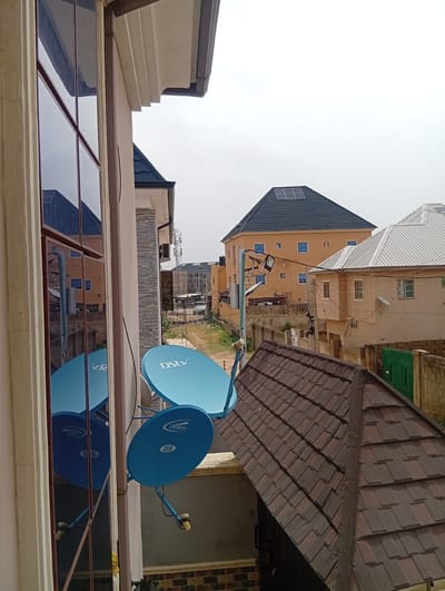 The image depicts an outdoor scene in Aviation Quarters, Nigeria, likely taken from an elevated position, such as a balcony or upper floor. The foreground contains two blue DStv satellite dishes mounted on a wall. A window reflects the buildings across the street and the satellite dishes themselves. A brown tiled roof is also in the foreground.

In the background, several residential buildings are visible. One has an orange facade with blue-trimmed windows and a black roof with solar panels installed. Adjacent to it is a beige building with a silver corrugated metal roof. A green door is visible on an outbuilding near a dilapidated stone wall. A road or walkway cuts through the middle of the scene, and several utility poles and power lines run between the buildings. The overall environment appears urban, with a mix of well-maintained and older structures. The sky is overcast, suggesting daytime.