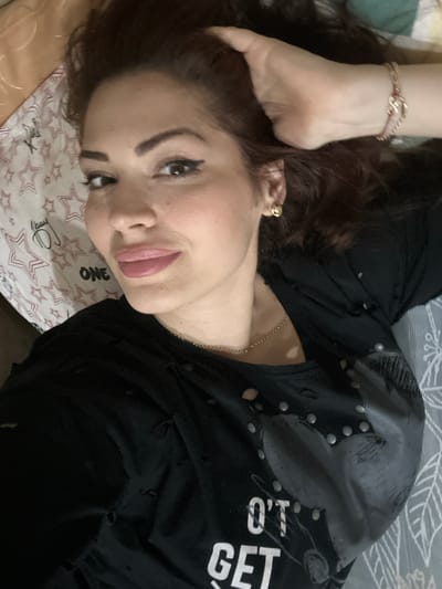 A woman is lying down, looking at the camera. She has dark hair and is wearing a black top with some silver accents. She is wearing an earring. Her hand is resting on her head. The background consists of bedding with a star pattern. The location is not possible to determine from this image.