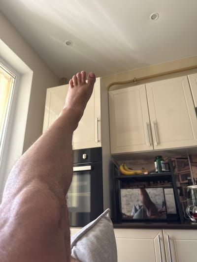 A first-person perspective shot shows a man's leg and foot extended upwards against a backdrop of a kitchen. The kitchen features cream-colored cabinets with silver handles, a black oven, a microwave with a reflective surface showing a reflection of the man, and a shelf with bananas and a green bottle. The ceiling has recessed lighting. The man's skin is fair and hairy. The scene is illuminated by natural light from a window on the left. The specific location is stated to be in the city of Romashkovo, Russia.