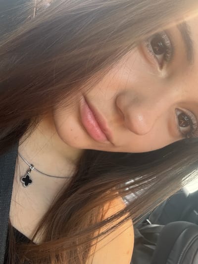 The image is a close-up selfie of a young woman's face, taken in Rakovski, Bulgaria. She has long, dark brown hair that partially obscures her face and shoulders. Her left eye is visible, with dark eyelashes. Her lips are slightly parted, and she is wearing a silver necklace with a black, clover-shaped pendant. The lighting suggests it is daytime, with sunlight casting a warm glow on her skin. The background is blurred but appears to be the interior of a vehicle, with dark upholstery visible. There are no other people or significant objects in the frame.