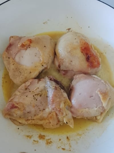 The image shows four pieces of cooked chicken thighs in a white pot or pan. The chicken pieces are arranged in a circular pattern, with their skins facing upwards. The skin is golden brown in some areas, indicating it has been seared or baked. There are visible seasonings, such as herbs, sprinkled on the chicken. A liquid, likely chicken broth or cooking juices, is pooled at the bottom of the pan. The lighting suggests an indoor setting, possibly a kitchen. No people are visible in the image. The focus is on the food, with the background being a simple white surface. The image does not provide any clues about the time of day, weather, or specific emotions. There is no visible text in the image. The location context of Oryol-Izumrud, Russia, does not offer any visual cues within the image itself.