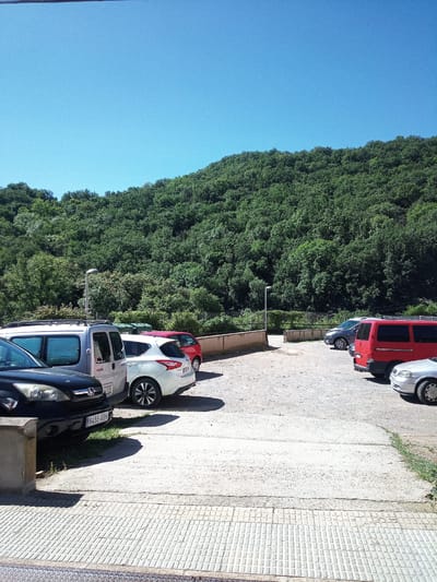 A sunny, clear day casts sharp shadows on a paved parking area at the base of a dense, tree-covered hillside in Castellfollit de la Roca, Spain. Several vehicles are parked, including a black SUV in the foreground on the left, a silver van next to it, a white hatchback, a dark blue sedan further back, and a red van and a silver car on the right side of the frame. The parking lot is gravelly with some patches of concrete. A single lamppost stands near the center of the image, behind the parked vehicles. A low, brown retaining wall runs along the edge of the parking lot. Green trash bins are visible behind the white hatchback. The hillside is covered in thick green foliage.