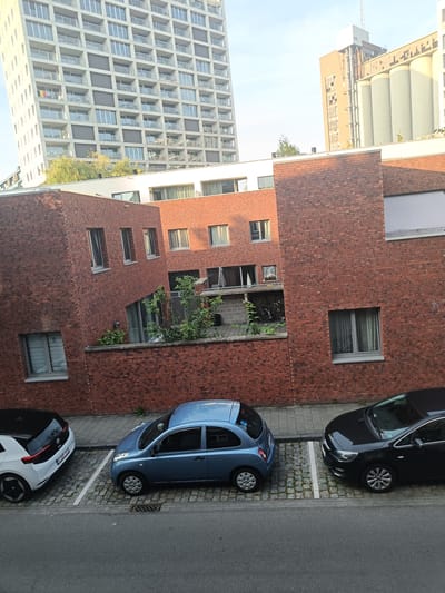 This image shows a street scene in Leuven, Belgium, featuring a multi-story modern apartment building and a brick building with multiple windows and balconies. In the foreground, there are three parked cars on a cobblestone parking area adjacent to the street. A white electric car with a black roof is parked on the left, followed by a small blue hatchback in the middle, and a black sedan on the right. The street itself is paved with asphalt. The brick building appears to have some greenery growing on balconies or in courtyards. The sky is visible, suggesting it is daytime.