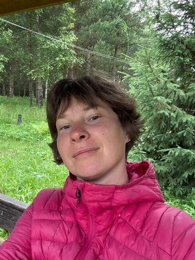 A person with short, brown hair and blue eyes is captured in a selfie. They are wearing a bright pink puffer jacket with a zipper. The individual is sitting outdoors, with a background of lush green grass and trees, including pine trees. A wooden bench is visible on the left side of the frame. The overall setting appears to be a natural, outdoor environment, possibly in or near Vorsma, Russia, given the mention of birch trees and the general landscape. The lighting suggests it is daytime, with soft natural light.