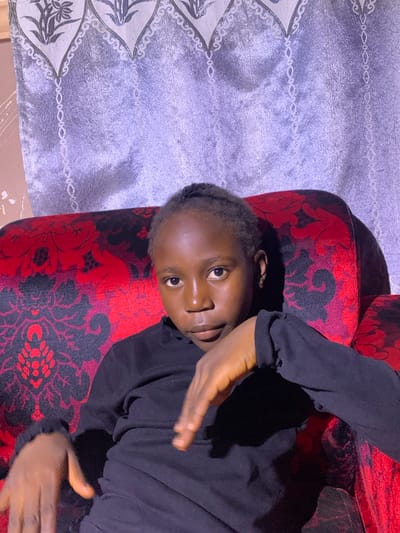A young Black person with braided hair is seated on a red and black patterned sofa. They are wearing a black long-sleeved shirt and are looking directly at the camera with their head tilted slightly. Their right arm is bent with their hand resting on their chest, and their left arm is extended downwards, with their hand open. The background consists of a silver and grey curtain with a floral pattern. The scene appears to be indoors, possibly in a living room. There is no specific indication of the location within Russo, Nigeria.