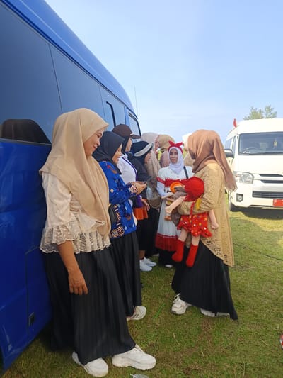 A group of women, predominantly wearing hijabs, stand alongside a blue passenger bus and a white van in an outdoor setting in Sukapura, Indonesia. The women are conversing and engaged in various activities. One woman holds a doll dressed in red. The surroundings include grass, suggesting a field or park-like area. The blue bus and white van indicate a transportation setting.