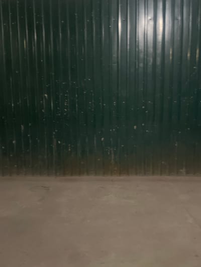 The image shows a close-up, head-on view of a dark green corrugated metal wall with a light grey concrete floor in the foreground. The wall has vertical ridges and appears to be slightly weathered with some light-colored specks scattered across its surface. The concrete floor is textured and appears dusty. The scene is devoid of any people or specific objects that would indicate a particular activity or location within Lebedinovka, Kyrgyzstan, beyond a generic industrial or utilitarian setting.