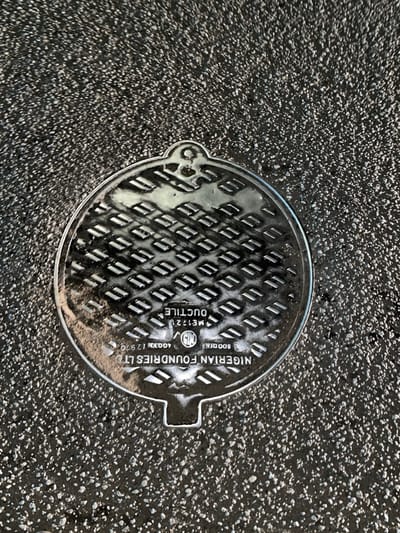 The image displays a circular metallic manhole cover embedded flush into a dark, granular paved surface, likely asphalt, in Babban Giji, Nigeria. The cover features a raised, repetitive diamond-plate pattern across its surface for grip. Inscribed on the cover are several details: "NIGERIAN FOUNDRIES LTD" is visible along the bottom curved edge. Above this, "800 DIA" (indicating diameter), "400KN I 2970," and "ME1221 DUCTILE" are legible, with "DUCTILE" inverted. A circular logo, possibly representing the Nigerian Standards Organisation (NISO) or similar, is present near the center bottom. The cover has a small, semi-circular extension with a circular hole at the top and a matching, solid semi-circular extension at the bottom. The surrounding pavement is uniform, dark gray with lighter aggregate specks. No people or other objects are present in the scene.