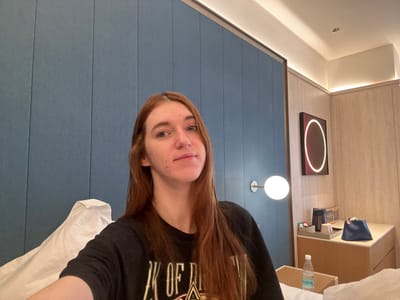 A selfie shows a fair-skinned woman with long, reddish-brown hair wearing a black t-shirt. She is positioned against a blue, textured wall. The background includes a modern hotel room with a nightstand featuring a water bottle and toiletries. An illuminated circular artwork hangs on a wood-paneled wall adjacent to a sphere-shaped wall light. Soft lighting illuminates the room. The perspective is slightly tilted.