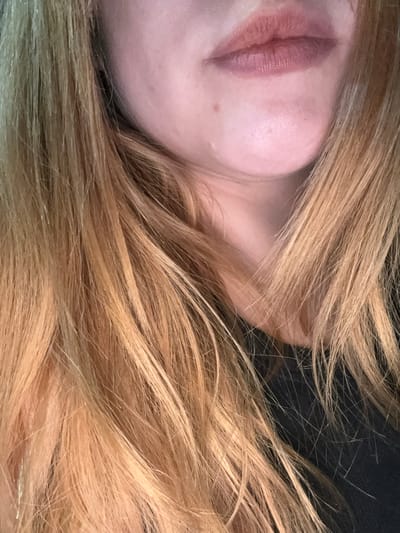 The image is a close-up, head-and-shoulders shot of a person with blonde hair and fair skin, focusing on their lower face and hair. Their lips are colored with a matte lipstick that appears to be a muted rosy-brown shade. A small mole is visible on the chin. The person is wearing a black garment, possibly a shirt or dress. The background is dark and out of focus, with strands of hair obscuring parts of it. There is no indication of the city of Getafe, Spain, or any other specific location.
