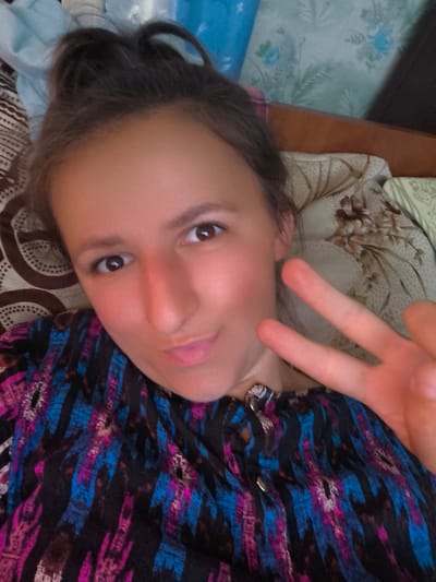A young woman with dark hair pulled up is lying down and taking a selfie. She is wearing a black shirt with a colorful abstract pattern of pink, blue, and white stripes. She is holding up two fingers in a peace sign with her right hand. Her left cheek is slightly flushed, and she has a slight smile on her face, with her lips puckered. The background appears to be a bed with patterned pillows and bedding. The lighting suggests it is daytime. The overall mood is casual and relaxed.