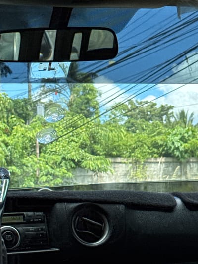 The image is taken from inside a vehicle, looking out through the windshield. The windshield has two circular suction cup marks on it, and there is a blue tinted film applied to the upper portion of the windshield, which also has a rearview mirror attached. Outside the vehicle, there is lush green foliage, a concrete wall, and a sky with some clouds and power lines visible. The interior of the vehicle includes a black dashboard with a car stereo and an air vent. This scene is likely located in Santa Cruz, Philippines, as indicated by the tropical vegetation.