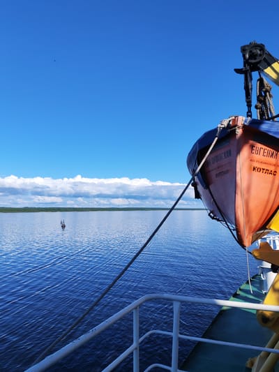 The image depicts a view from a boat on a river or large body of water. A lifeboat, painted orange and blue, is secured on the boat's side. Text in Russian is visible on the lifeboat. A small navigation buoy marks the waterway. On the horizon, a line of trees borders the water, with a band of clouds in the sky above. The sky is largely clear and blue. The location might be near the city of Kotlas, as that is written on the lifeboat. Prislon' is not directly identified but could be nearby the area where this photo was taken. The general scene suggests calm weather and daytime.