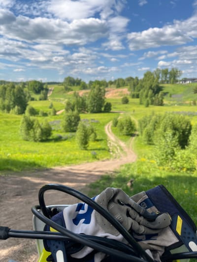 This image, captured in Новочувашский, Russia, presents a rural landscape from the perspective of an individual positioned on a dirt bike or similar off-road vehicle. In the immediate foreground, the black handlebars and control cables of a vehicle are prominently displayed. Beneath the handlebars, a white and blue motocross garment is visible, partially obscured by a pair of grey and black motocross gloves, which feature a visible Husqvarna logo on the wrist strap. The gloves are resting on the garment and a dark blue component of the vehicle's dashboard or front fairing.

The midground reveals a vast, undulating green landscape characterized by several unpaved dirt tracks winding through grassy fields. Numerous deciduous trees and bushes are scattered across the terrain, with some areas showing patches of purple wildflowers, likely lupines, amidst the green and occasional yellow flora. The dirt tracks suggest a prepared area for off-road riding.

In the background, the landscape continues with rolling green hills and a dense treeline under a bright blue sky. Numerous white, fluffy cumulus clouds are distributed across the sky. On the far right horizon, indistinct structures, possibly houses or buildings, are faintly visible. The scene is illuminated by strong daylight, indicating clear weather conditions. No individuals are visible in the frame.