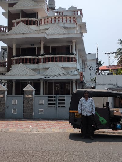The scene depicts a daytime street view in Kochi, Kerala, India, featuring a multi-story building, an auto-rickshaw, and a man.
The prominent structure on the left is a four-story building, predominantly white with reddish-brown architectural accents. It features multiple balconies with terracotta-colored balustrades and supporting columns. The ground floor entrance includes a grey metal pedestrian gate with square vents and a matching larger vehicle gate with horizontal slats, flanked by a grey stone or tiled wall. A vertical red sign with white text, partially obscured, is mounted on the building's right side, displaying characters suggestive of "HOME STAY." The building's flat roof supports several white cylindrical water tanks and a white railing.
Positioned on the street level to the right of the building is a black auto-rickshaw, identified by its Kerala license plate "KL 07 M 2027." The auto-rickshaw has yellow trim along its canopy and a bright green guitar graphic painted on the passenger side's fabric cover.
Standing next to the auto-rickshaw, facing the viewer, is an adult male. He has a dark complexion, dark hair, and a mustache. He wears a light-colored, long-sleeved button-up shirt with a dark, intricate pattern, dark trousers, and dark shoes. His right hand is in his pocket.
The foreground consists of a dark asphalt road. A sidewalk with reddish-brown and lighter-colored interlocking pavers separates the road from the building's frontage. The background, to the right of the auto-rickshaw, shows a lighter-colored building with a visible red-tiled roof section, utility poles, and sparse green foliage. The sky is clear and bright.