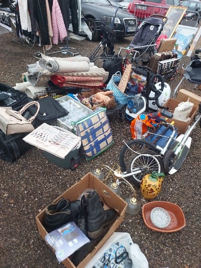 The image depicts an outdoor market stall in Eckbolsheim, France. The ground is covered with dark gravel. The scene is densely packed with various used goods for sale.
In the foreground, a brown cardboard box contains several pairs of dark boots and other packaged items. Adjacent to this box, a brass-colored chandelier with three frosted glass shades, a yellow ceramic jug with floral patterns, and a reddish-brown rectangular dish holding a clear, patterned glass plate are displayed on the ground.
To the left of the center, a large stack of rolled rugs and textiles, primarily in neutral and earth tones, rests on the ground. Nearby, a beige handbag, two black handbags, and a black backpack are placed. A white, flat object, possibly a book or sign with text and images, lies on top of a dark green container. A blue and white patterned tote bag is positioned next to it.
The mid-ground features a variety of items: a plastic doll with curly brown hair lying face-up amidst a collection of small objects, a light blue woven basket, an orange toy weapon, a white helmet, and various electronic components or small appliances. Further back, a dark-colored stroller is visible, along with a bicycle wheel and other mechanical parts. Two framed artworks, one a dark painting and another a lighter landscape print, lean against other items.
In the background, several vehicles are parked, including a dark grey Audi sedan with a partial French license plate (VV-435-FS) and a red vehicle. Clothing on hangers is visible on portable racks to the far left. No individuals are discernible within the frame interacting with the merchandise.